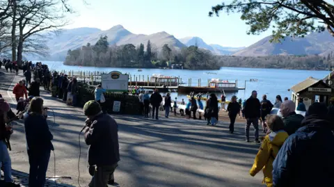 Ian Gordon Shot of individuals standing on the edge of a lake with mountains in the background. The water is bright blue as well as the sky. People are wearing thick jackets and hats. 