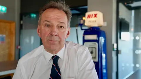 Duncan Lambert, wearing a white shirt and a blue tie sits in a room in front of an old Rix Petroleum petrol pump