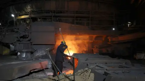 A worker in a hard hat stokes the furnace, which is bright orange. There are steel objects around the man.