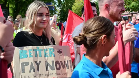 Pacemaker A young woman with long blonde hair holds a brown cardboard sign which says 'better pay now, fair pay to guard your day'. She is wearing a black t-shirt and a silver cross necklace. There are other men and women surrounding her holding red union flags. They are wearing red, blue, white and other coloured clothing.