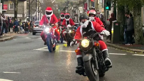 A group of five bikers in Santa suits watched by crowds.