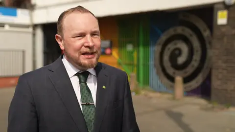 A fair bearded man in a suit jacket and green and blue patterned tie and tie clip stands in front of a rainbow-coloured gate on a street 