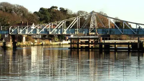 Evelyn Simak/Geograph A railway bridge can be seen over a river, with its reflection visible in the water on a sunny day