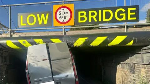 The back of a silver van which has become stuck under a bridge. The back doors have been wedged against the bridge - which has bright yellow stripes and yellow letters reading, Low bridge - 2.0m - 6'-6''.