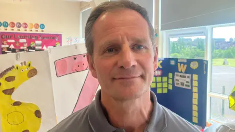 Robert is looking into the camera. He's wearing a grey top and standing in a classroom in a primary school. Children's artwork is hanging behind him.