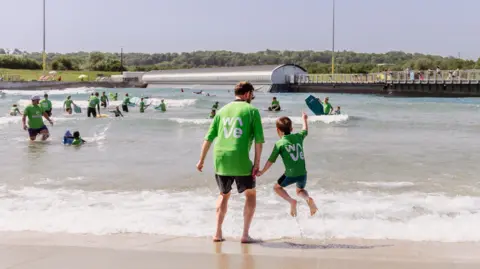 The Wave A man holding hands with a young boy as they jump over the ongoing waves which are lapping at the short. In the background, there are lots of other surfers wearing green rash vests, playing in the waves and swimming with body boards.
