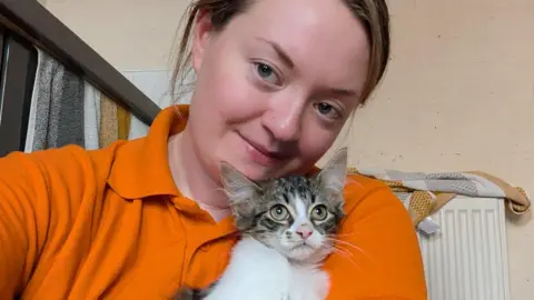 A woman with brown hair pulled back behind her head and wearing an orange top, has a cat just beneath her chin, looking off camera. Behind them is a radiator with a towel on top of it.