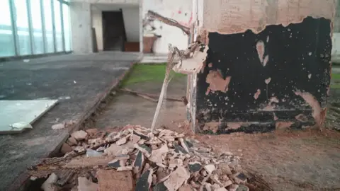 The picture shows the inside of a building that is being stripped out or undergoing heavy refurbishment. Rubble is piled on the floor in the foreground, made up of broken plaster, tiles and chunks of concrete. A damaged column stands nearby, with its outer surface chipped away so that metal mesh and fragments of plaster are left hanging loose.