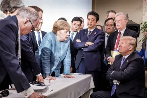 Getty Images The then-German Chancellor Angela Merkel leans over a table to speak to a seated Donald Trump during a G7 summit in 2018 in Canada. They are surrounded by world leaders including Theresa May, UK prime minister, Emmanuel Macron, French president, Shinzo Abe, Japan prime minister, and John Bolton, US national security adviser.