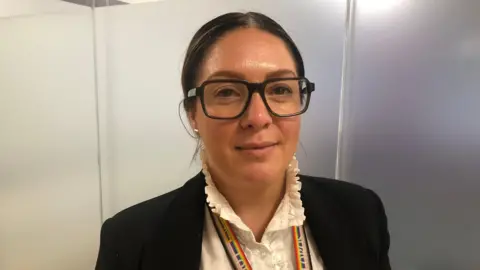 Camilla Barrow wearing black-framed glasses and a white collared top and black jacket. 
