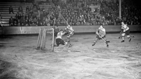 Beamish, the Living Museum of the North A hockey game at Durham Ice Rink in 1961. The audience is looking on as players are skating. They chasing the puck and attempting to score.