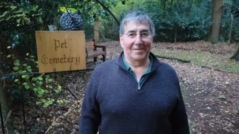 A man in a black fleece. He is standing in a wooded pet cemetery. 