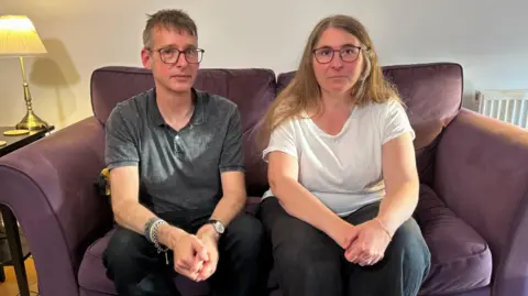 Chris, a man with glasses and wearing a grey polo shirt, is sat on a purple sofa next to his wife Diana, who had long light brown hair and is wearing glasses and a white T-shirt. They are both looking at the camera