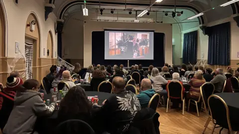 People sit around round tables on black chairs with gold trim. They look up at a large screen showing a film.