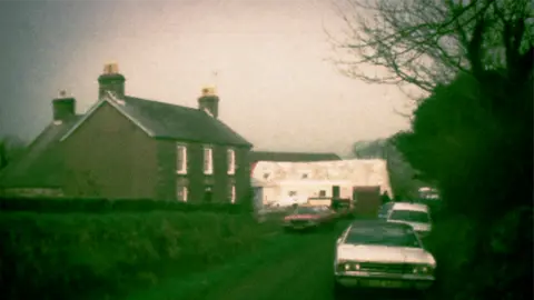 BBC News Archive representation  from 1976 of a grey farmhouse connected  a lane, with a achromatic  barn adjacent  to it and tons  of aged  constabulary  cars parked outside