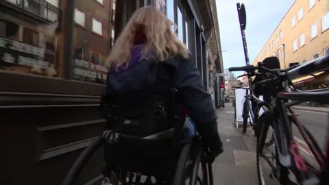 Abbi Brown wheeling her wheelchair on a narrow pavement in between shopfronts, bike racks and a-boards on a London street