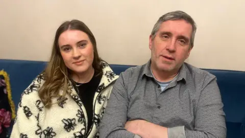 Family handout Aimee sitting on a sofa with her dad Damien. Aimee has long brown hair with blonde ends, she's wearing a black top and a cream zip up cardigan with black flowers. Damien has light grey short hair and is wearing a grey shirt with a light blue top underneath.