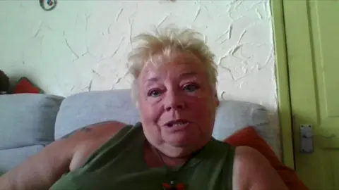 A woman wearing a green vest top sits on a grey sofa in a living room. 