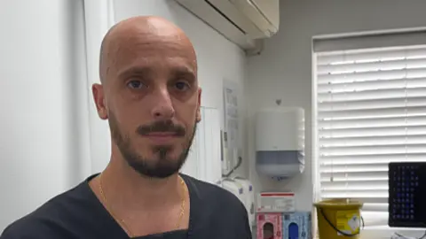 Dr Dario Sales, who has a shaved head with a brown beard and moustache, is photographed wearing a black tunic in his dental surgery.