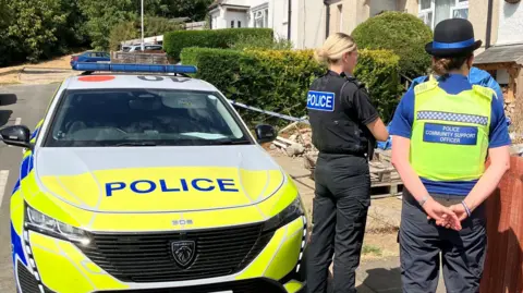 A female police officer is standing next to a police community support officer in a street. They are next to a police car and facing a row of houses.