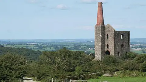 A rural landscape shows an old stone engine house with a tall chimney standing on a hillside beside a winding dirt path, fields, and low stone walls stretching across the countryside.