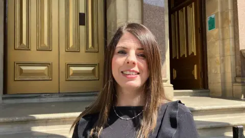 Anne Marie Gallagher, who has long brown hair, stands in front of a pair of gold doors, she is wearing a black dress