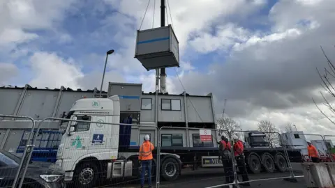 A crane with a white cab is using chains to grab a grey metal cabin above a complex of grey cabins, arranged in two stories. Three people in orange hi-vis jackets and white hard hats are looking upwards to watch the movement. There is metal fencing in the foreground. The sky is cloudy.