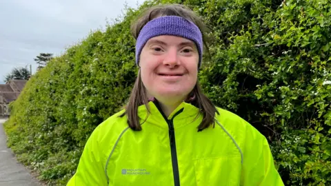 A woman in a lime green running jacket and a purple headband stood in front of a green hedge