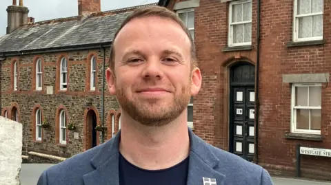 Man in dark blue t-shirt and light blue jacket with a ginger beard standing in front of a row of houses