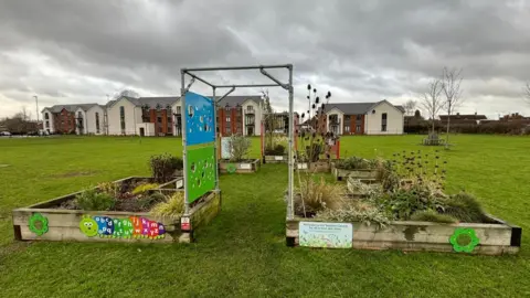 Prestbury Parish Council The sensory garden, set in a large green space with flats visible in the background. There are a number of large raised planters and a metal archway. 