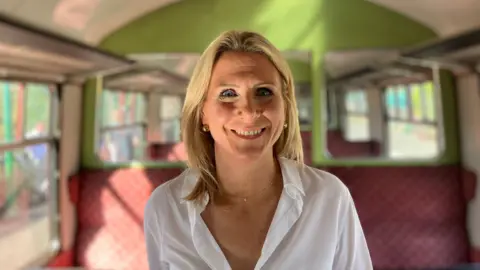Henry Godfrey-Evans/BBC Erica Gilson has blonde shoulder-length hair and is wearing a white open-necked shirt. She is standing in the vintage train carriage, and is grinning.