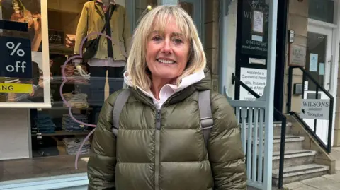 A woman with blonde hair and a dark green puffer coat, wearing cream mittens and smiling outside a shop in Ilkley.