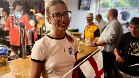 Churchdown Club A woman with blonde hair is stood at the bar of a pub wearing a white England shirt and holding an England flag