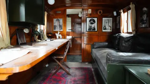 PA Media The inside of a caravan which has wooden stained walls. There is a dark green sofa on the right and a large table on the left which has a wooden chair tucked under it. There are several pictures and items hanging on the wall.