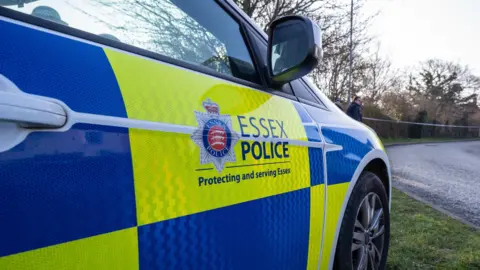 BBC A blue and yellow police car parked on the side of a rural road. Essex Police is written on its door, with the Essex Police crest.