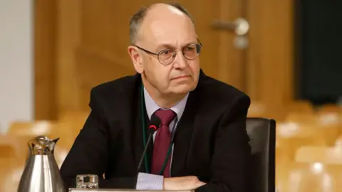 Scottish Parliament A man with receding grey hair and glasses sits with his arms folded at a desk. he is wearing a dark suit, blue striped shirt and burgundy tie. 