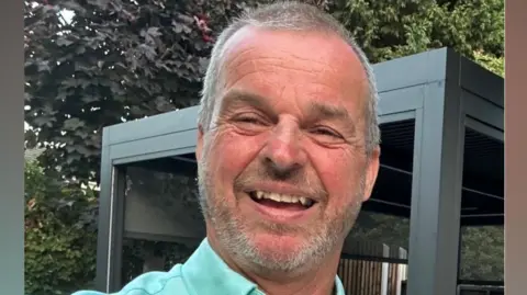 A selfie of Mark Beech who is smiling and has grey hair and facial hair. He is wearing a bright teal blue shirt and is in a garden with trees in the background