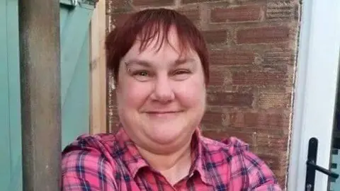 Jane wearing a pink and blue checked shirt smiling with a brick wall and pale green door behind her