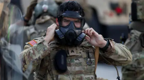 California National Guard member in camouflage colored uniform puts on black gas mask
