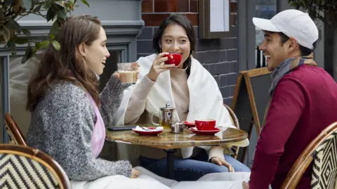 Two women and a man sit at an outdoor table drinking coffee.