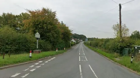Google A Google street view of a rural single-carriageway road with trees and hedges either side. Houses can be seen in the distance.