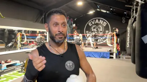 A man waiting a black vest is stood in front of a boxing ring and looking towards the right of the camera. He is holding a boxing glove in his left hand and the words 'Evans boxing fitness' is painted on the wall behind the boxing green.