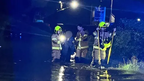 An older woman wades through ankle deep water flanked by two firefighters