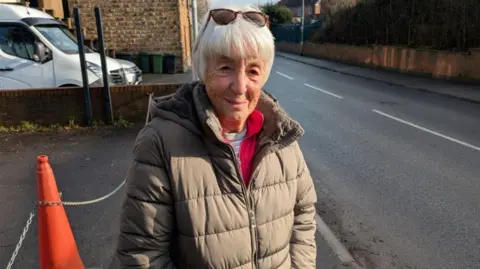David Spereall/BBC A woman standing on a suburban road. She has white hair and is wearing sunglasses on her head. She is wearing a grey-coloured coat that is bathed in sunlight.