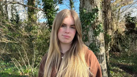 A young woman with long blonde hair who is wearing a brown sweatshirt
