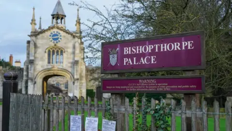 Getty Images Entrance gate to to Bishopthorpe Palace, the home of the Archbishop of York, includes a stone gatehouse with a clock in the distance with a purple sign stating the name of the building and the coat of arms of the Archbishop