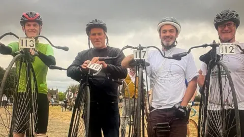 Lee Stanbury Participants stand with penny farthing bicycles at an outdoor event ahead of a ride.
