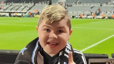 Family handout Alfie standing in a football stadium. He has blonde hair and is giving a thumb's up. He's wearing a white and black scarf and football top.