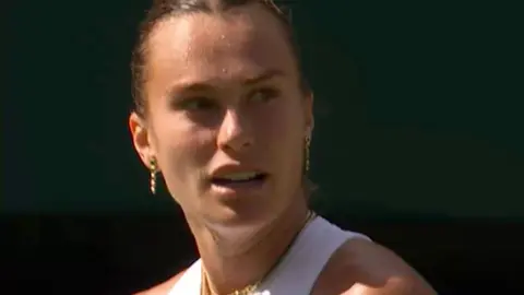 Aryna Sabalenka looks to the right - she has brown hair pulled back and wears a white vest top