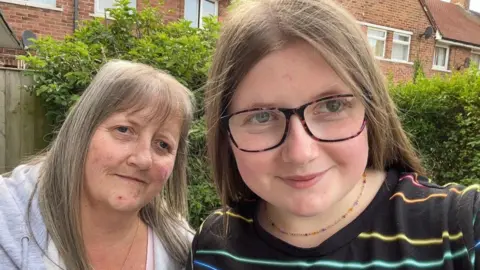 BBC Ffion, wearing a striped t-shirt and her mum Lorraine, wearing a grey hoodie, look in the camera and smile. In the background, there are some bushes from their garden.